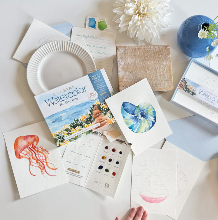 A table displays Schiffer Publishing’s "Watercolor the Easy Way Paint-Your-Own Watercolor Cards Coastal" kit, with paint samples, brushes, blank and painted notecards (jellyfish, seashell), a palette, and a white flower in a blue vase.