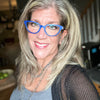 A smiling woman with highlighted hair wears bright blue glasses and a silver chain necklace, standing indoors near a staircase. She’s the perfect example for Schiffer Publishing, Ltd.'s "Projects and Techniques for Metal Clay Jewelry.