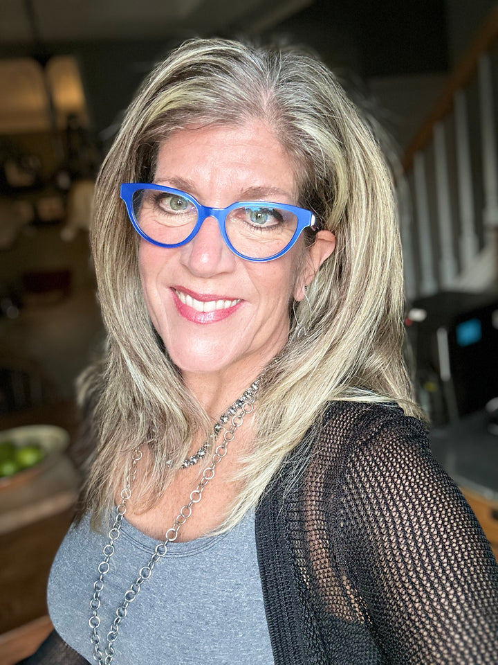 A smiling woman with highlighted hair wears bright blue glasses and a silver chain necklace, standing indoors near a staircase. She’s the perfect example for Schiffer Publishing, Ltd.'s "Projects and Techniques for Metal Clay Jewelry.