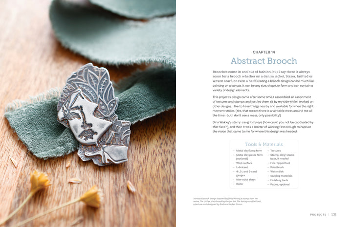 A silver abstract brooch with engraved leaves and a face sits on green fabric beside "Projects and Techniques for Metal Clay Jewelry" from Schiffer Publishing, Ltd., showcasing detailed jewelry craftsmanship.