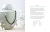 A silver pendant necklace with green beads rests on a pale green ceramic container. In the softly blurred background is Projects and Techniques for Metal Clay Jewelry by Schiffer Publishing, Ltd., open to a page about silver jewelry.