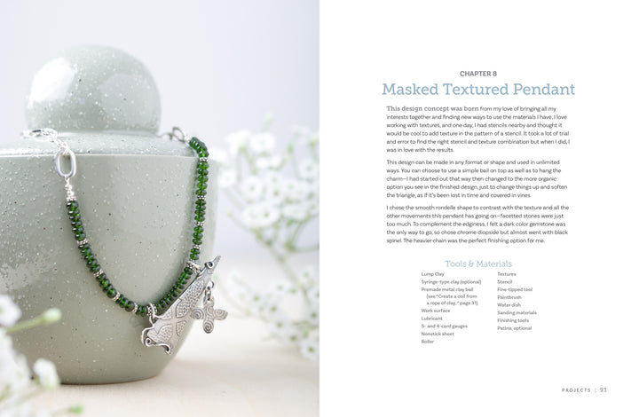 A silver pendant necklace with green beads rests on a pale green ceramic container. In the softly blurred background is Projects and Techniques for Metal Clay Jewelry by Schiffer Publishing, Ltd., open to a page about silver jewelry.
