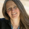 A woman with long brown hair and blue eyes smiles softly, wearing a "Fiber Craft Heritage" shirt by Schiffer Publishing, Ltd., dark jacket, and silver necklace. The blurred background features soft earthy tones.