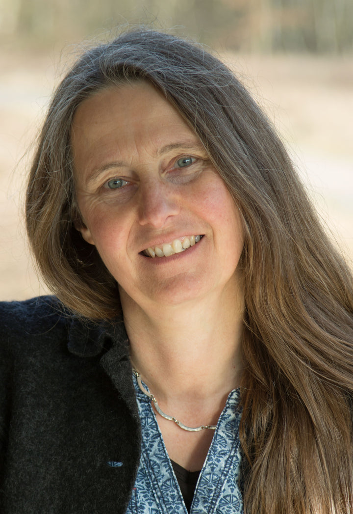 A woman with long brown hair and blue eyes smiles softly, wearing a "Fiber Craft Heritage" shirt by Schiffer Publishing, Ltd., dark jacket, and silver necklace. The blurred background features soft earthy tones.