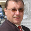 A man in a suit, red tie, and round sunglasses smiles by a fountain. With short brown hair and a mustache, he looks ready to share tips from "Green Wood Turning" by Schiffer Publishing, Ltd. for woodturning beginners.