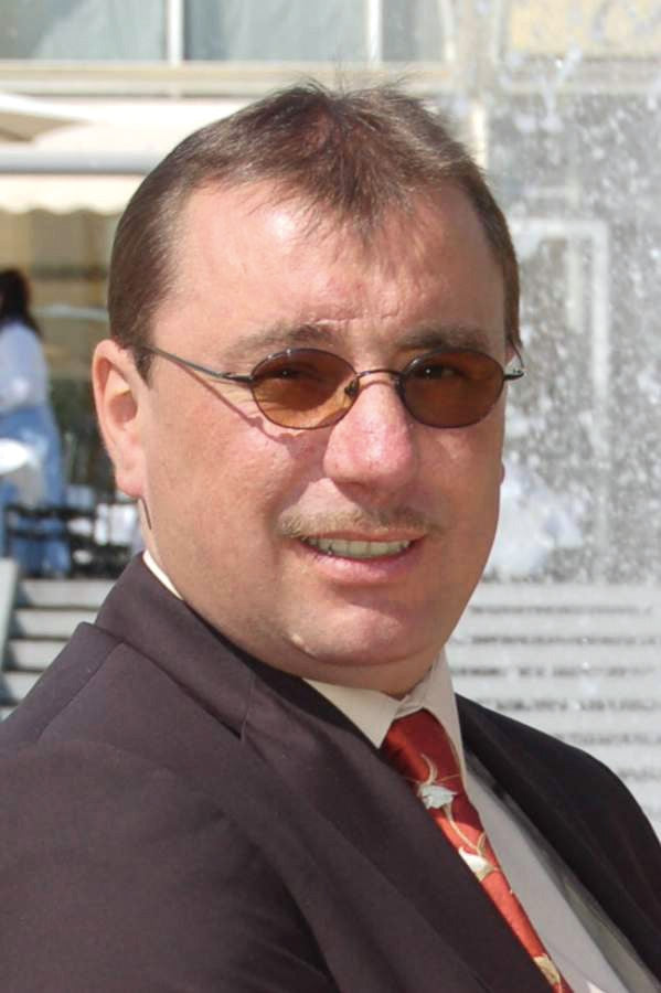 A man in a suit, red tie, and round sunglasses smiles by a fountain. With short brown hair and a mustache, he looks ready to share tips from "Green Wood Turning" by Schiffer Publishing, Ltd. for woodturning beginners.