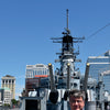 A man in a blue shirt stands before the US Coast Guard Cutter 37 by Schiffer Publishing, Ltd., a historic warship docked among other ships in Baltimore, with city buildings and a clear blue sky in the background.