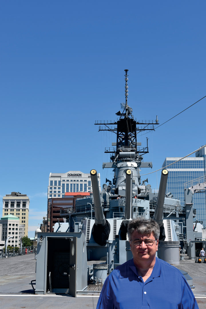 A man in a blue shirt stands before the US Coast Guard Cutter 37 by Schiffer Publishing, Ltd., a historic warship docked among other ships in Baltimore, with city buildings and a clear blue sky in the background.