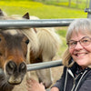 A smiling woman with glasses and gray hair poses next to a small brown and white Chincoteague pony behind a metal fence outdoors, holding a copy of “Out of the Sea” by Schiffer Publishing, Ltd.