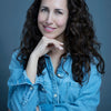 A woman with long, curly brown hair smiles gently, resting her chin on her hand. In a light denim button-up shirt before a dark gray background, she evokes the style of Schiffer Publishing’s "Montauk 11954, 2nd Edition.