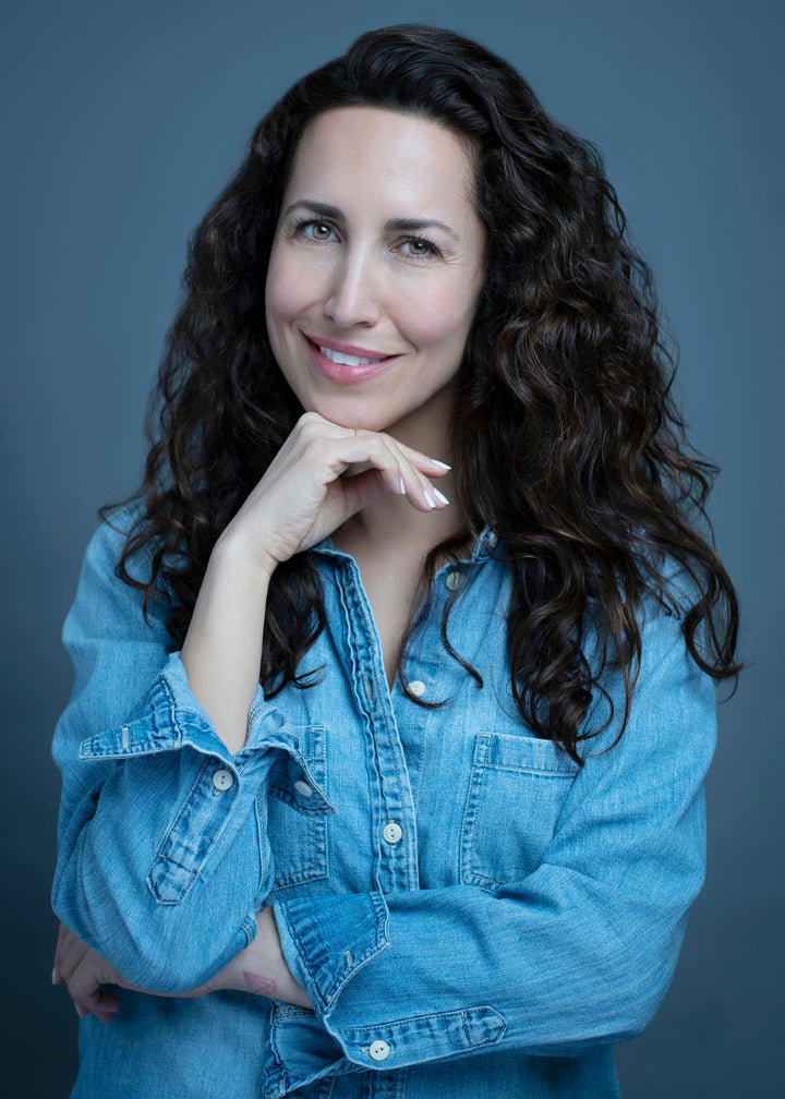 A woman with long, curly brown hair smiles gently, resting her chin on her hand. In a light denim button-up shirt before a dark gray background, she evokes the style of Schiffer Publishing’s "Montauk 11954, 2nd Edition.