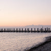 A serene Montauk 11954 waterfront sunset with a wooden pier, pebble beach, and white building—perfect for any photography collection. Featured in "Montauk 11954, 2nd Edition" by Schiffer Publishing, Ltd., in soft pink and orange hues.