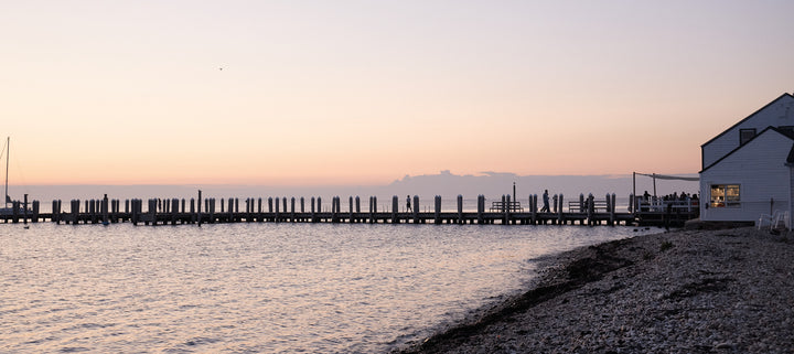 A serene Montauk 11954 waterfront sunset with a wooden pier, pebble beach, and white building—perfect for any photography collection. Featured in "Montauk 11954, 2nd Edition" by Schiffer Publishing, Ltd., in soft pink and orange hues.