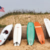Surfboards of various colors and sizes stand in sand before grassy dunes and an American flag—a classic Montauk scene featured in Schiffer Publishing, Ltd.’s "Montauk 11954, 2nd Edition.