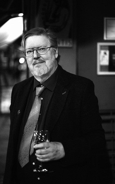 A man with glasses, beard, and mustache stands indoors in a suit and tie, holding a wine glass. The black-and-white image evokes the mysterious atmosphere of The John Dee Oracle from Schiffer Publishing, Ltd.