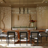 A dining room inspired by Penny Drue Baird, as featured in "PowerHouse" by Schiffer Publishing, Ltd., with a wooden table, eight upholstered chairs, a floral mural, glass chandelier, chevron floor, and a side table with a vase. Light streams in from the left.