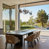 The PowerHouse dining room by Schiffer Publishing, Ltd. showcases beige chairs and a dark wood table, large glass doors to a sunny patio, pool, garden with mountain views, and geometric pendant lights.