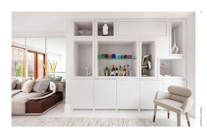 A stylish living area with a built-in white shelving unit displays colorful décor—perfect inspiration from PowerHouse by Schiffer Publishing, Ltd. A beige armchair is up front; a sofa with pillows sits by the large windows on the left.