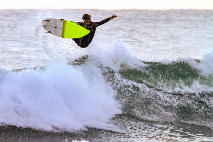 A surfer in a wetsuit launches above the wave, yellow and white board airborne—embodying the thrill of discovering uncharted breaks, as seen in surfEXPLORE by Schiffer Publishing, Ltd., with ocean spray all around.
