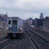 A graffiti-covered subway car travels on a weathered elevated track past city apartments, evoking NYC history as seen in "New York Subways and Stations, 1970–1990" from Schiffer Publishing, Ltd.