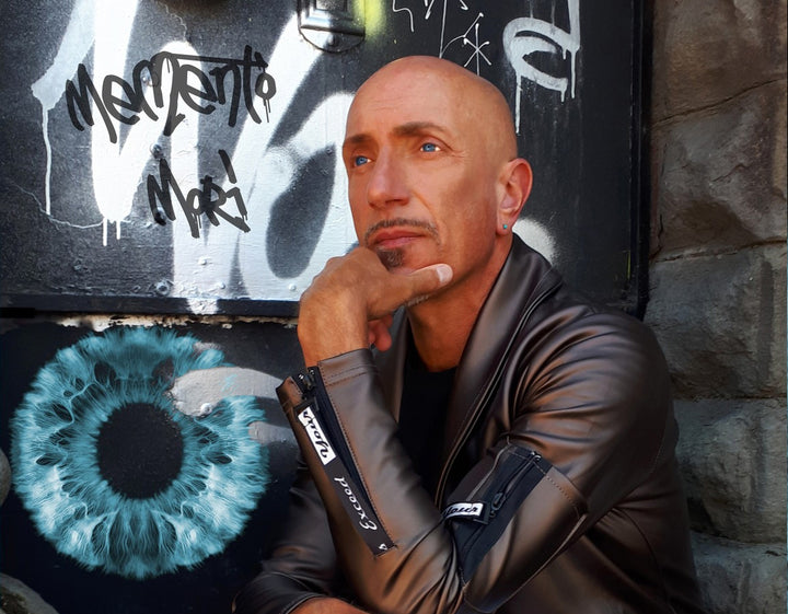 A bald man in a leather jacket sits thoughtfully before a graffiti wall and painted blue eye, evoking the vivid, surreal style of the Starman Tarot Remastered by Schiffer Publishing, Ltd., inspired by David Bowie.