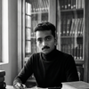 A man in a dark turtleneck sits at a desk, writing about unexplained mysteries with Morbid Kuriosity’s by Schiffer Publishing, Ltd., surrounded by stacks of books and a filled bookshelf, all in a black and white atmosphere.