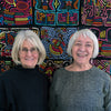 Two older women with gray hair and glasses smile in front of a vibrant, geometric fiber art textile, as featured in "Contemporary Basketry" by Schiffer Publishing, Ltd.
