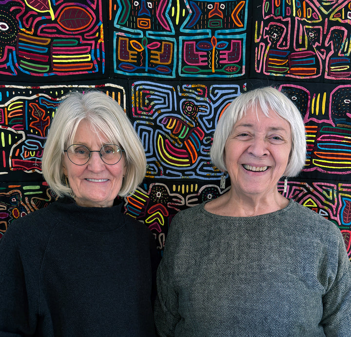 Two older women with gray hair and glasses smile in front of a vibrant, geometric fiber art textile, as featured in "Contemporary Basketry" by Schiffer Publishing, Ltd.