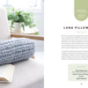 A beige armchair holds an open book and a chunky knit gray pillow pouf, inspired by "Get Started in Finger Knitting" from Schiffer Publishing, Ltd., basking in soft natural light with a cream blanket draped over the chair’s back.