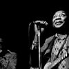 A Black man sings and plays guitar on stage, capturing the spirit of American music, while another musician plays harmonica in the dimly lit scene. “Inside the Moment” by Schiffer Publishing, Ltd. celebrates this musical artistry.