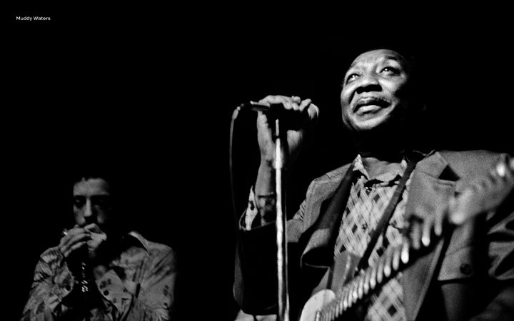 A Black man sings and plays guitar on stage, capturing the spirit of American music, while another musician plays harmonica in the dimly lit scene. “Inside the Moment” by Schiffer Publishing, Ltd. celebrates this musical artistry.