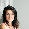 A woman with long dark hair and fair skin sits against a light gray backdrop in a white sleeveless top, softly lit by window light, evoking the mystical feel of The Ukrainian Tarot by Schiffer Publishing, Ltd.