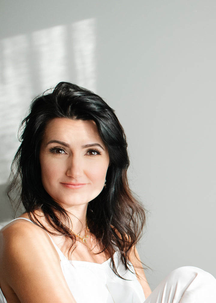 A woman with long dark hair and fair skin sits against a light gray backdrop in a white sleeveless top, softly lit by window light, evoking the mystical feel of The Ukrainian Tarot by Schiffer Publishing, Ltd.