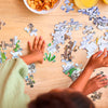 Two children assemble The Tiniest Giant 4-in-1 Puzzle Set by Schiffer Publishing, Ltd. on a wooden table covered with puzzle pieces, with snacks and juice on the side.