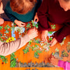 Four people collaborate on The Tiniest Giant 4-in-1 Puzzle Set by Schiffer Publishing, Ltd., arranging green and floral pieces at a wooden table. The overhead photo shows only their hands and arms, ideal for fans of The Tiniest Giant.