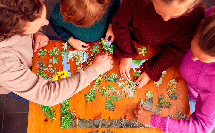 Four people collaborate on The Tiniest Giant 4-in-1 Puzzle Set by Schiffer Publishing, Ltd., arranging green and floral pieces at a wooden table. The overhead photo shows only their hands and arms, ideal for fans of The Tiniest Giant.