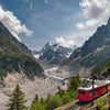 A red train from Alpine Railways by Schiffer Publishing, Ltd. winds through lush mountains, glacier views, steep peaks, and green forests—showcasing one of the most scenic European Alps routes under a partly cloudy sky.