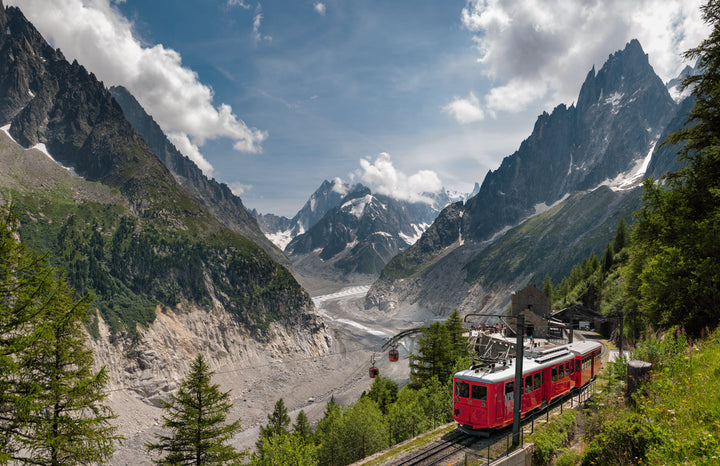 A red train from Alpine Railways by Schiffer Publishing, Ltd. winds through lush mountains, glacier views, steep peaks, and green forests—showcasing one of the most scenic European Alps routes under a partly cloudy sky.