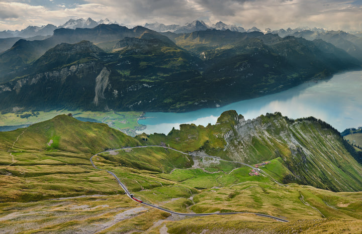 A panoramic view of winding mountain roads and green hills above a blue lake evokes the dramatic scenery featured in "Alpine Railways" by Schiffer Publishing, Ltd., reminiscent of classic European Alps train journeys.