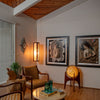 A cozy mid-century modern living room features a wooden ceiling, tan chairs, two portraits of women, and a glowing Modeline of California pendant light by Schiffer Publishing, Ltd., adding warmth above a table with green glassware.