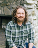 A bearded man with curly brown hair sits outdoors on a rustic stone wall, smiling at the camera. He is on The Law of One Contemplation Deck by Schiffer Publishing, Ltd., wearing a green plaid shirt and evoking themes from the deck.