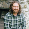 A bearded man with curly brown hair sits outdoors on a rustic stone wall, smiling at the camera. He is on The Law of One Contemplation Deck by Schiffer Publishing, Ltd., wearing a green plaid shirt and evoking themes from the deck.