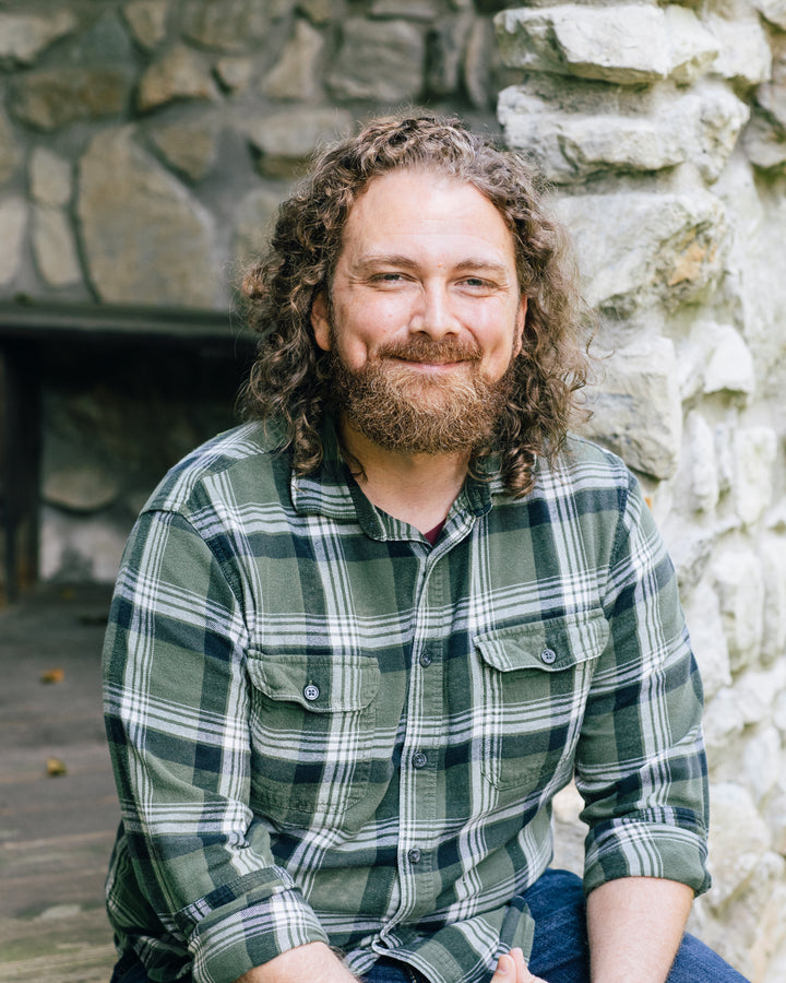 A bearded man with curly brown hair sits outdoors on a rustic stone wall, smiling at the camera. He is on The Law of One Contemplation Deck by Schiffer Publishing, Ltd., wearing a green plaid shirt and evoking themes from the deck.