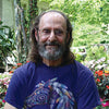 An older man with long hair, a beard, and glasses in a colorful horse t-shirt smiles in a lush garden, perhaps reflecting on quotes from The Law of One Contemplation Deck by Schiffer Publishing, Ltd. among blooming flowers on a sunny day.