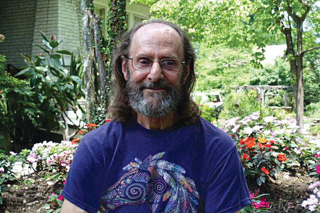 An older man with long hair, a beard, and glasses in a colorful horse t-shirt smiles in a lush garden, perhaps reflecting on quotes from The Law of One Contemplation Deck by Schiffer Publishing, Ltd. among blooming flowers on a sunny day.