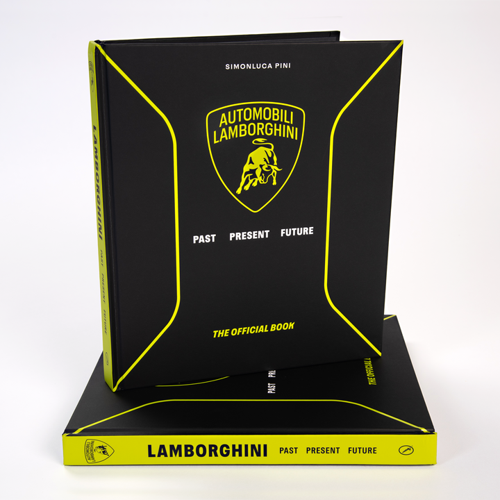 A black and yellow book titled "Automobili Lamborghini" by Schiffer Publishing, Ltd. stands upright on another copy, featuring a sleek cover with the iconic logo—a perfect gift for fans of luxury sports cars and Lamborghini legends.