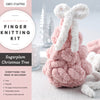 A pink, textured, handcrafted Christmas tree with a white yarn bow sits on a table beside the Super Simple Finger Knitting Kit Sugarplum Christmas Tree by Schiffer Publishing, Ltd., showing its included items.