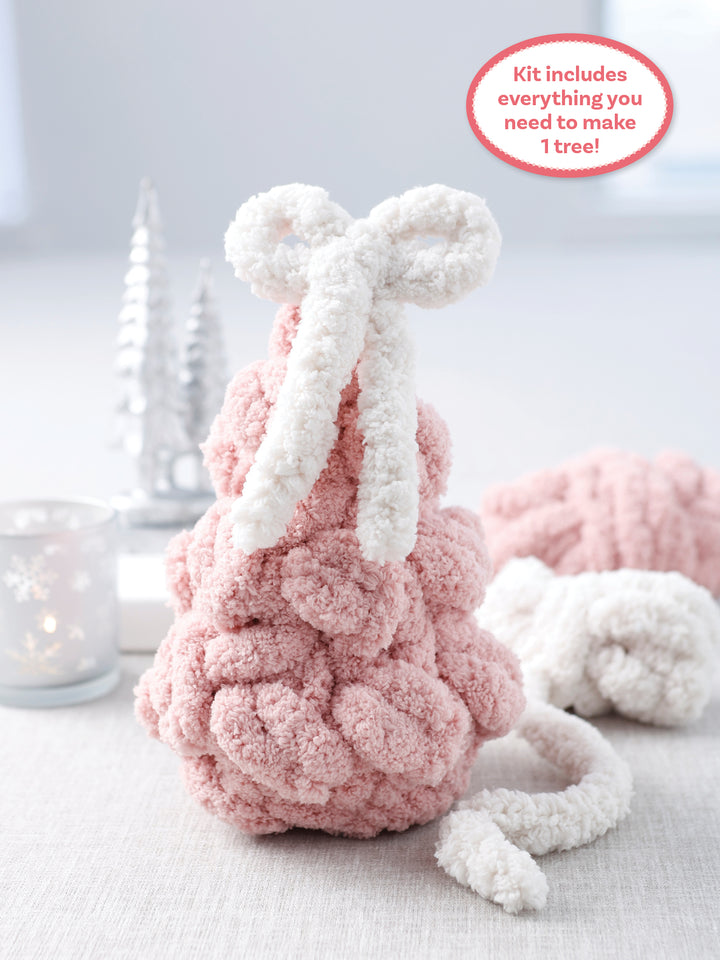 The Super Simple Finger Knitting Kit Sugarplum Christmas Tree by Schiffer Publishing, Ltd. sits on a table with a big white bow, surrounded by a snowflake candle holder and two silver trees in the background.