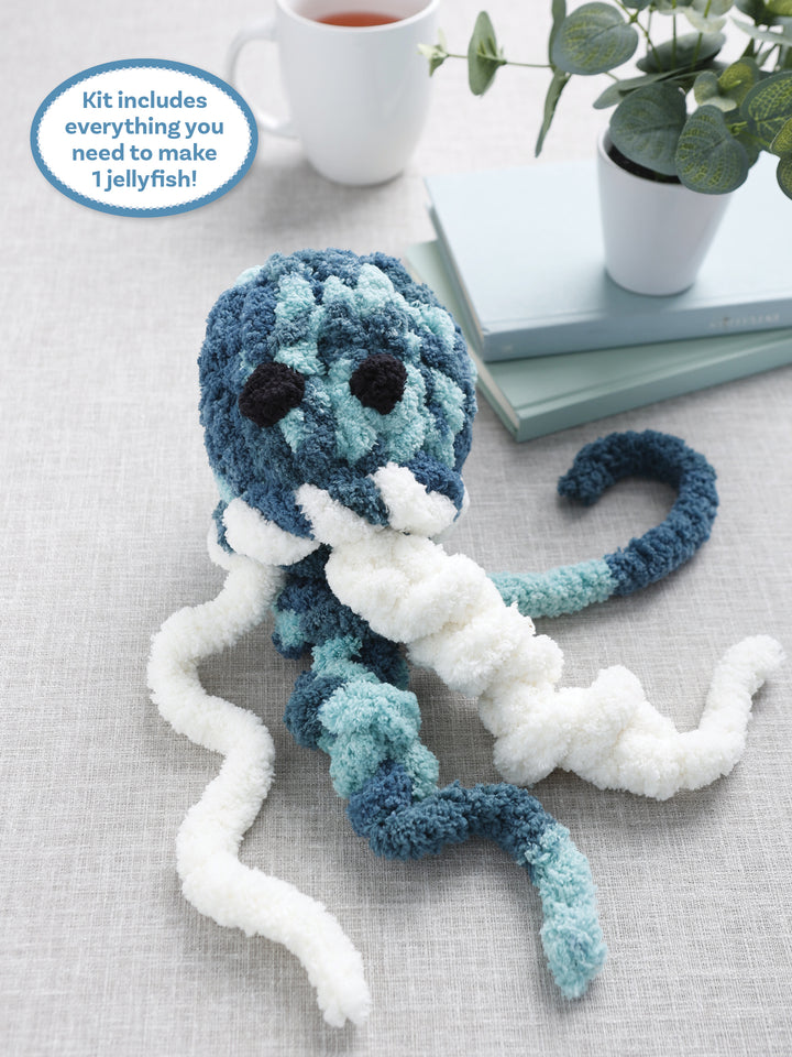 A plush blue and white jellyfish with big black eyes sits by books and tea. The label: Super Simple Finger Knitting Kit Jiggle the Jellyfish by Schiffer Publishing, Ltd.—includes everything needed to make your own cute jellyfish!.