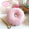 The Crochet Cafe Amigurumi Kit Donut by Schiffer Publishing, Ltd. features a pink crocheted donut with light blue sprinkles on a white surface beside a gold spoon and blurred crochet supplies in the background. Text highlights its beginner focus.